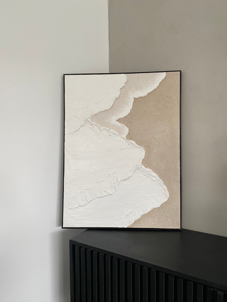May include: Abstract art painting in a black frame with white and beige textured paint. The painting is hanging on a wall above a black cabinet.