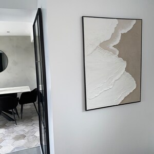 May include: A white and beige abstract painting with a textured surface, framed in black, hanging on a white wall.