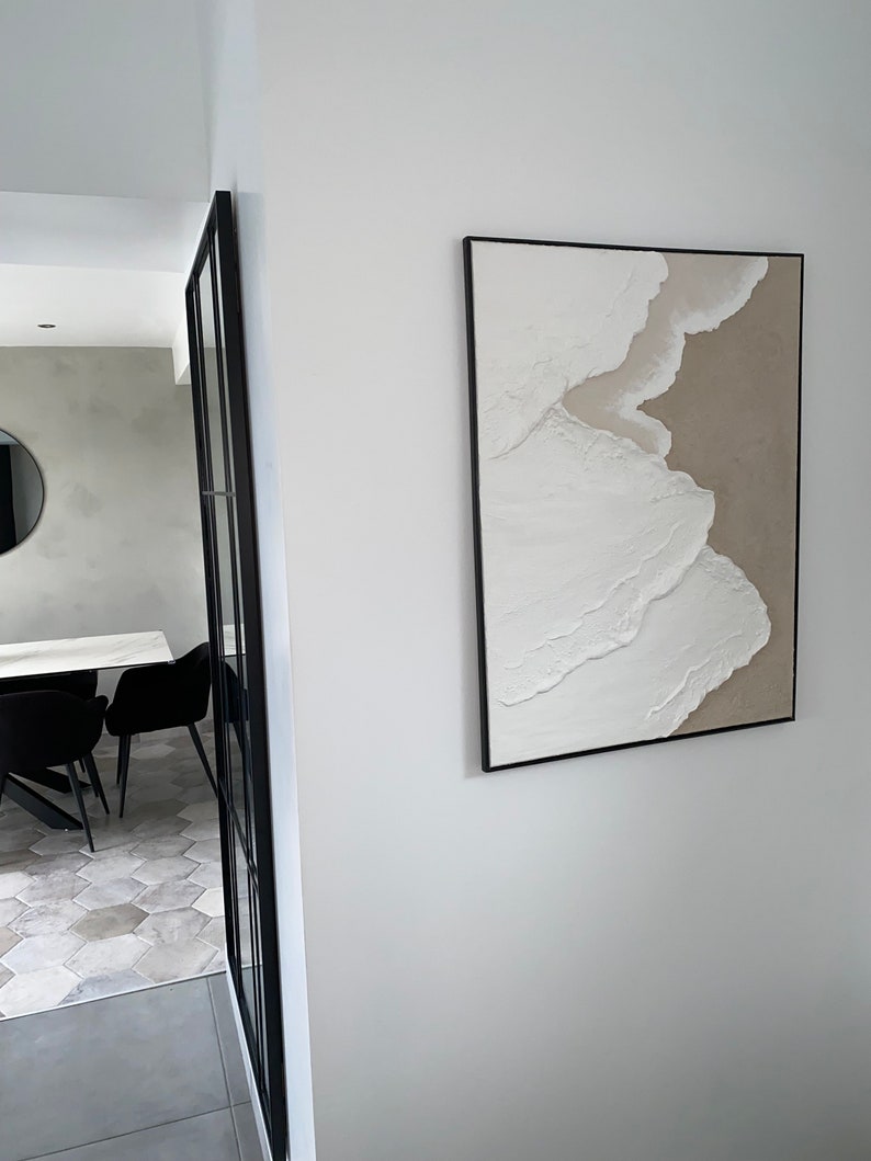May include: A white and beige abstract painting with a textured surface, framed in black, hanging on a white wall.