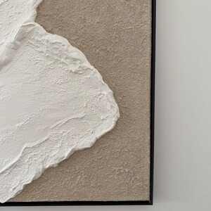 May include: Abstract painting with a textured white shape on a beige background. The painting is framed in black.