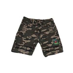 Puede incluir: Pantalones cortos de mezclilla con estampado de camuflaje, acabado desgastado y roturas deshilachadas en los muslos. Incluyen paneles acanalados sobre las rodillas, costuras horizontales negras y un parche bordado de una rana en la pierna.