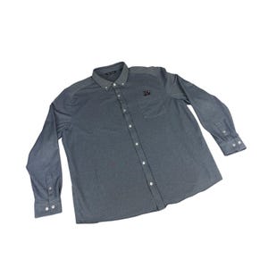 Peut inclure: Une chemise grise boutonnée à manches longues, avec un col et une poche poitrine. La chemise a un petit emblème sur la poche et est faite d'un tissu doux et texturé. La chemise est boutonnée et semble neuve.