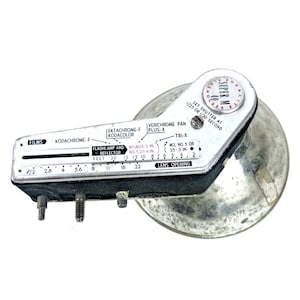May include: Vintage Super M flash unit with a silver reflector and a black and silver control panel. The panel features settings for films, lens openings, and shutter speeds. The text "SUPER M" is visible.