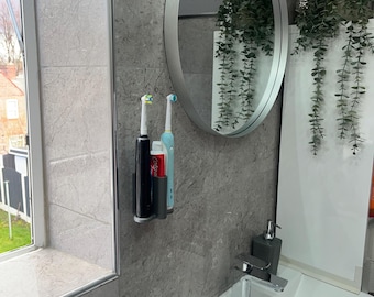 Puede incluir: Un organizador de baño de pared gris oscuro que sostiene dos cepillos de dientes eléctricos y un tubo de pasta de dientes Colgate. El organizador está colocado sobre una pared de azulejos grises junto a un espejo redondo, un lavabo blanco y un grifo cromado.