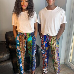 Puede incluir: Dos personas con camisetas blancas y pantalones de estampado colorido. Los pantalones están hechos de diferentes telas y tienen un diseño de patchwork. Las personas están de pie frente a una pared gris.
