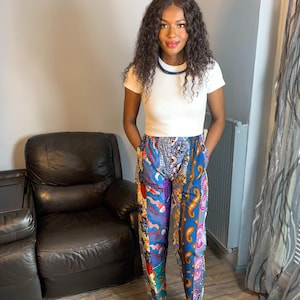 May include: A woman wearing a white t-shirt and colorful patterned pants. She is standing in front of a leather chair and a patterned curtain.