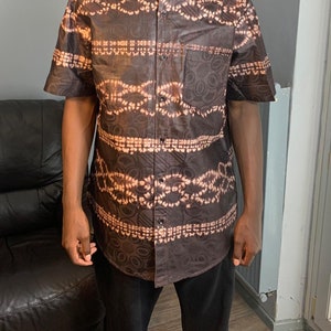 Puede incluir: Una camisa de manga corta marrón con un diseño estampado. La camisa tiene un cuello mandarín y cierre de botones.