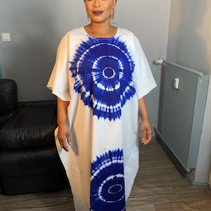 Peut inclure: Une robe maxi blanche avec un motif tie-dye bleu. La robe a un col rond et des manches courtes. Le motif présente deux grands cercles avec un cercle plus petit au centre.