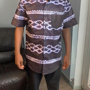 Puede incluir: Una camisa de botones de manga corta con estampado negro y blanco con un diseño tie-dye. La camisa tiene un bolsillo en el lado izquierdo.