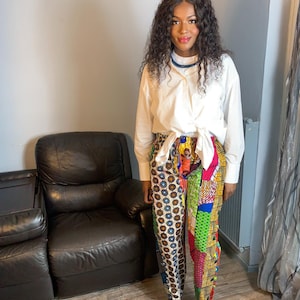 May include: A woman wearing a white button-down shirt tied at the waist and colorful patterned pants. She is wearing black sandals and has long curly hair.