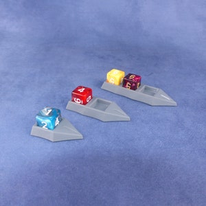 Op de afbeelding: Drie grijze plastic dobbelstenenbakken met verhoogde randen, elk met een dobbelsteen in een andere kleur. De dobbelstenen zijn rood, geel en blauw met witte cijfers.