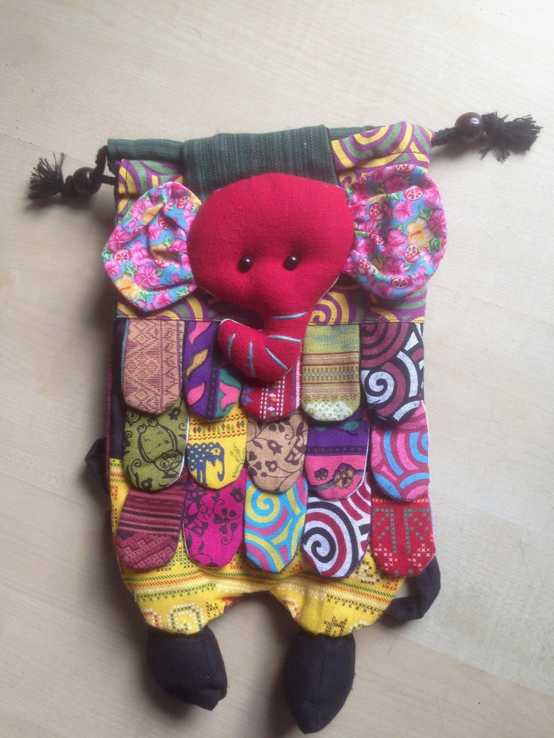 Pu&ograve; includere: Una borsa a cordoncino a forma di elefante in patchwork colorato con una testa di elefante in feltro rosso e piedi in feltro nero. La borsa &egrave; realizzata con diversi tessuti con motivi e colori diversi, tra cui rosa, viola, giallo, verde e blu.