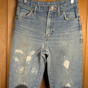 May include: Light blue denim jeans with white paint splatters and dark blue patches on the knees. The jeans are hanging on a wooden hanger. The jeans have a button closure and classic five-pocket design.