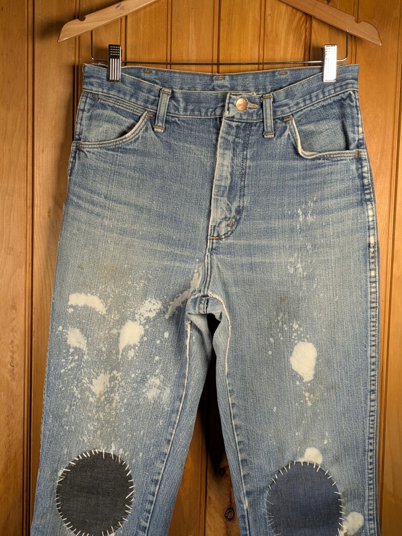 May include: Light blue denim jeans with white paint splatters and dark blue patches on the knees. The jeans are hanging on a wooden hanger. The jeans have a button closure and classic five-pocket design.