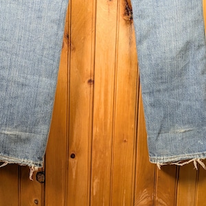 May include: Light blue denim jeans with a slight flare at the bottom. The jeans have a distressed look with frayed edges at the hem and a worn patch on the left leg. The background is a wooden wall.