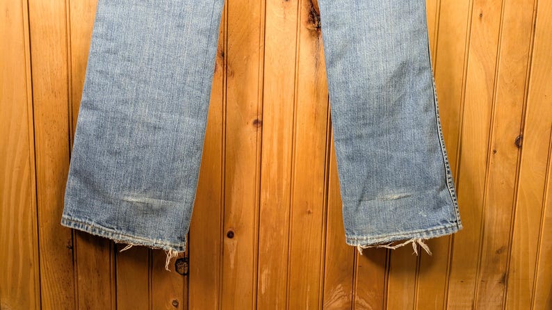 May include: Light blue denim jeans with a slight flare at the bottom. The jeans have a distressed look with frayed edges at the hem and a worn patch on the left leg. The background is a wooden wall.