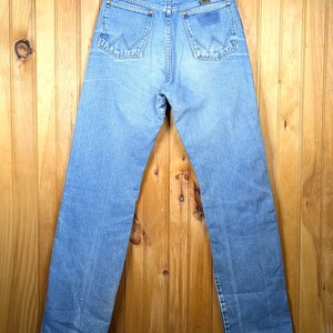 May include: Light blue denim jeans with the Wrangler logo on the back pocket. The jeans are hanging on a wooden hanger against a wooden background. The jeans have a classic five-pocket design and a straight leg.