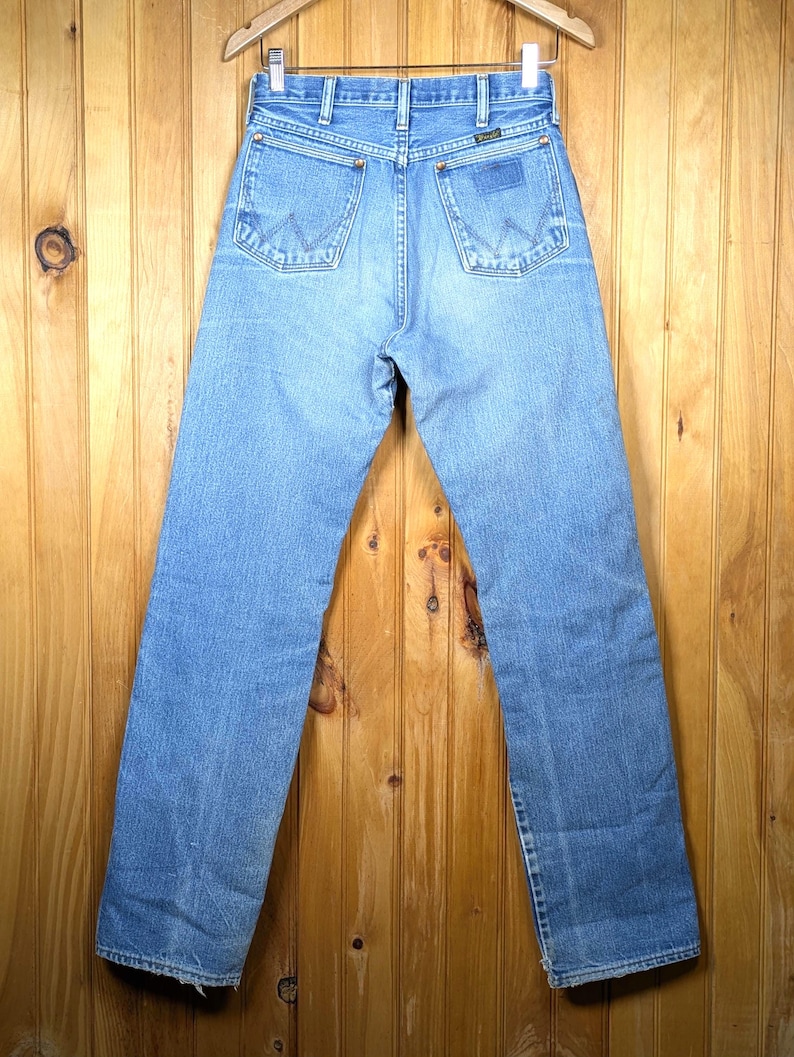 May include: Light blue denim jeans with the Wrangler logo on the back pocket. The jeans are hanging on a wooden hanger against a wooden background. The jeans have a classic five-pocket design and a straight leg.