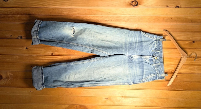 May include: Light blue denim jeans with a distressed knee detail, folded cuffs, and a button closure. The jeans are laid flat on a wooden surface with a wooden hanger.