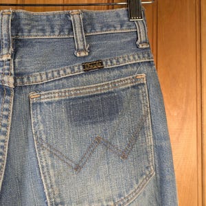 May include: Close-up of light blue denim jeans with a Wrangler back pocket. The pocket features a stitched 'W' design and is set against a faded denim backdrop. The jeans are hanging on a wooden hanger.