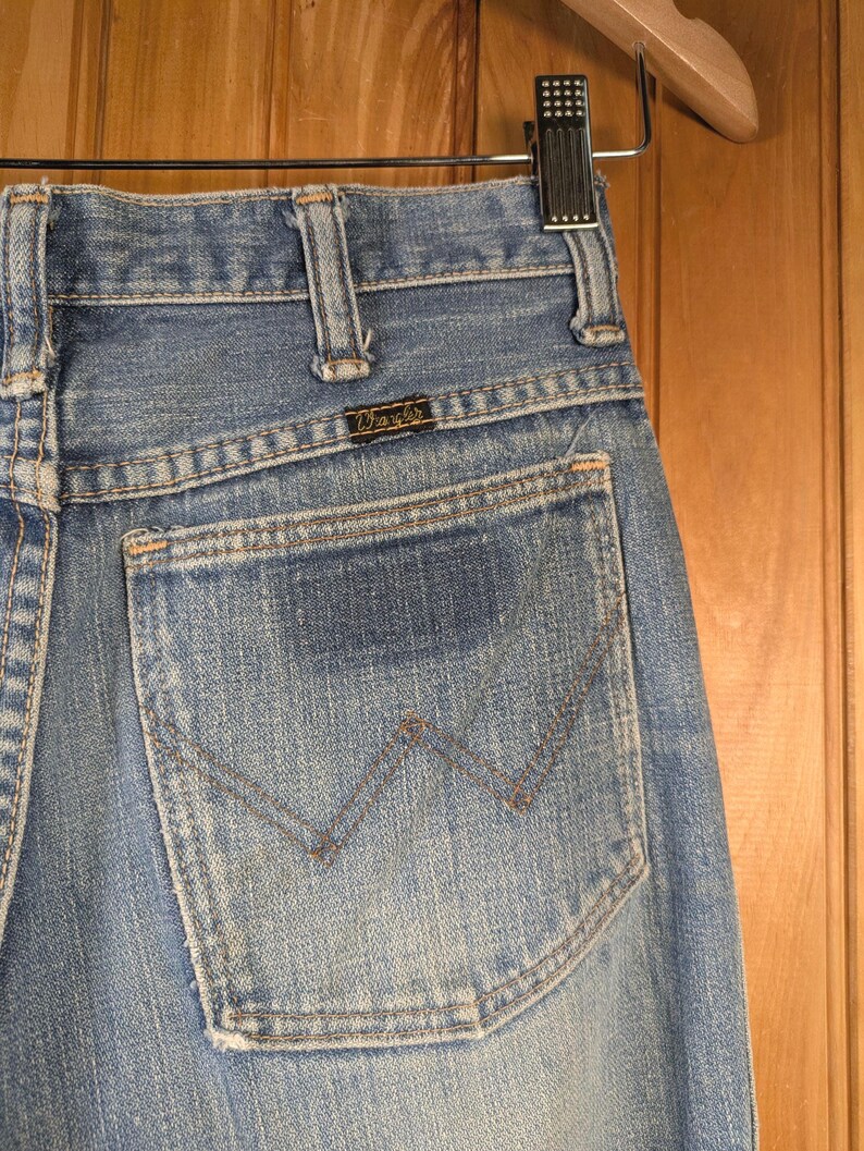 May include: Close-up of light blue denim jeans with a Wrangler back pocket. The pocket features a stitched 'W' design and is set against a faded denim backdrop. The jeans are hanging on a wooden hanger.