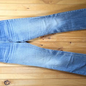 Op de afbeelding: Lichtblauwe denim jeans met een klassiek vijf-zakken ontwerp, gepresenteerd op een houten kleerhanger. De jeans hebben een vervaagde wassing en liggen plat op een houten oppervlak, wat hun rechte pijpen en algehele stijl benadrukt.