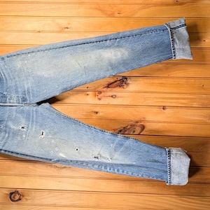 May include: Light blue denim jeans with distressed details, folded cuffs, and a wooden hanger. The jeans are laid flat on a wooden surface, showcasing their faded wash and worn appearance. A classic casual wear item.