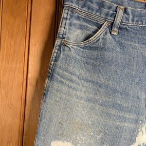 May include: Close-up of light blue denim jeans with white bleach-like splatters. The jeans have a classic five-pocket design and are hanging on a wooden hanger. The background is a wooden wall.