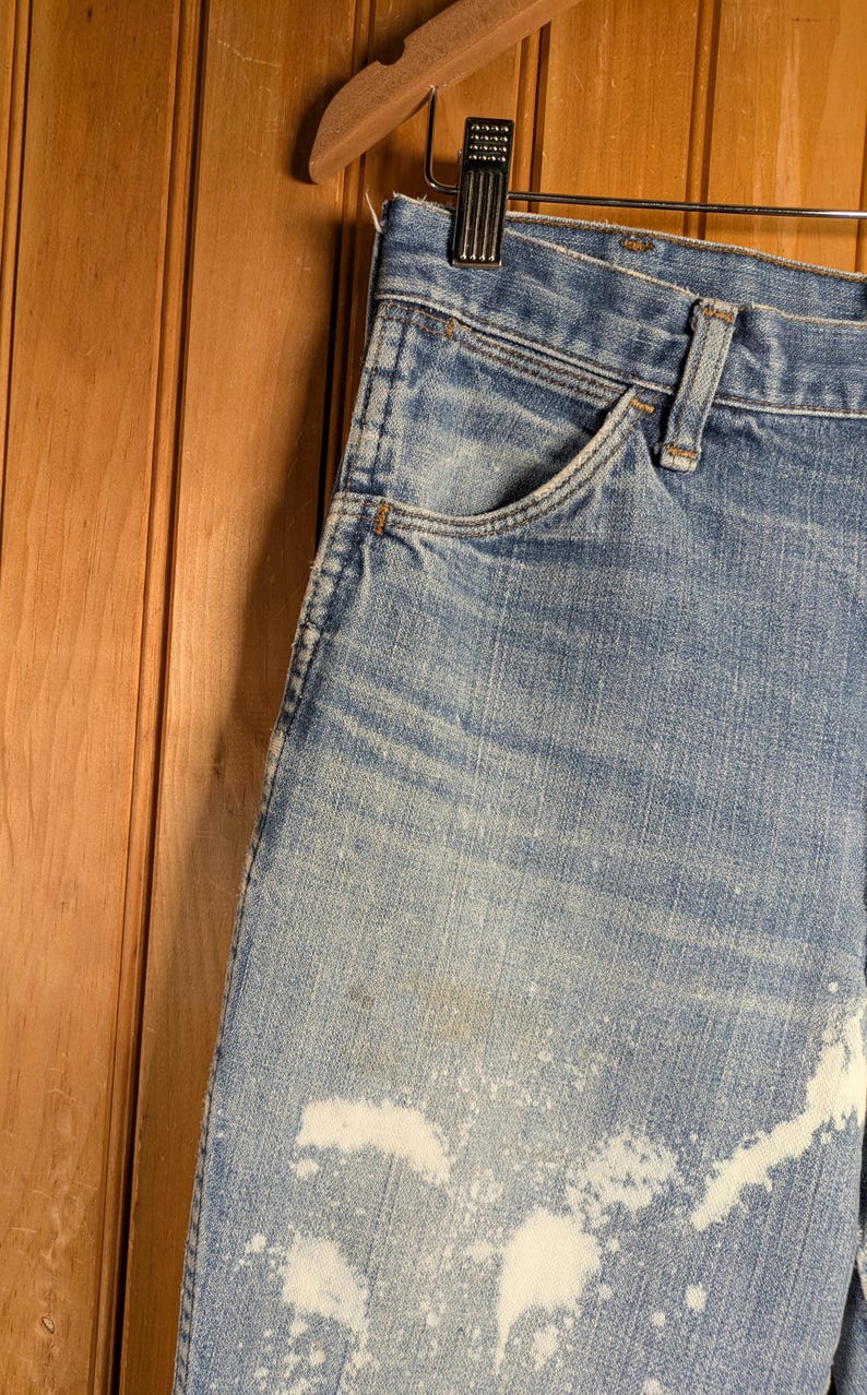 May include: Close-up of light blue denim jeans with white bleach-like splatters. The jeans have a classic five-pocket design and are hanging on a wooden hanger. The background is a wooden wall.