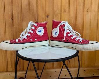 May include: A pair of red high-top canvas sneakers with white laces, white rubber toe caps, and white rubber soles featuring a black stripe. Each shoe displays a circular white patch on the inner ankle with the blue text Converse All Star.
