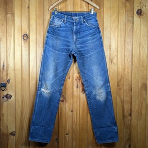May include: Blue denim jeans with a distressed look, hanging on a wooden hanger. The jeans have a faded appearance with a tear at the knee and light wear marks. The background is a wooden wall.