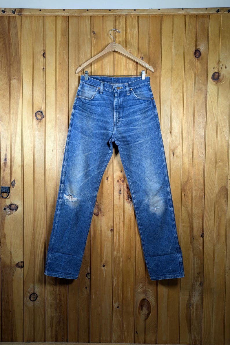 May include: Blue denim jeans with a distressed look, hanging on a wooden hanger. The jeans have a faded appearance with a tear at the knee and light wear marks. The background is a wooden wall.