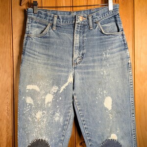 May include: Light blue denim jeans with white paint splatters and dark blue patches on the knees. The jeans are hanging on a wooden hanger against a wood-paneled background. The jeans have a button fly and classic five-pocket design.