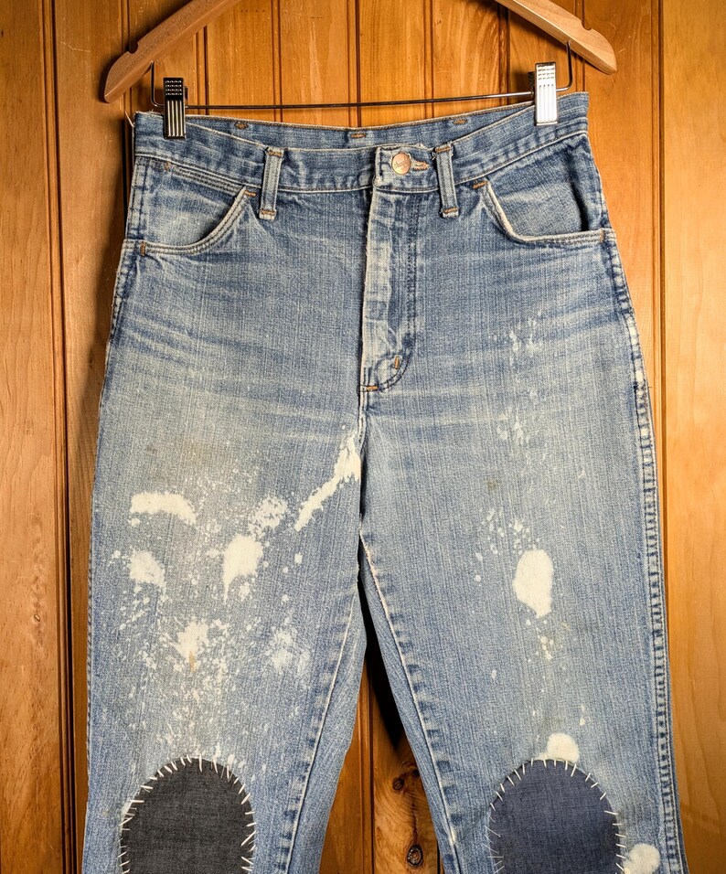 May include: Light blue denim jeans with white paint splatters and dark blue patches on the knees. The jeans are hanging on a wooden hanger against a wood-paneled background. The jeans have a button fly and classic five-pocket design.