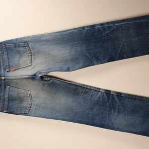 May include: Blue denim jeans with a faded wash, visible back pockets, and a leather patch. The jeans are hanging on a wooden hanger against a neutral background. The jeans have a classic, straight-leg fit.