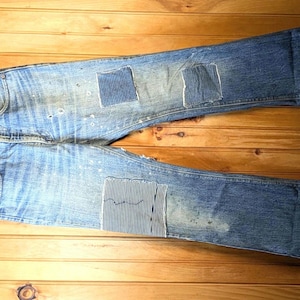 Pode incluir: Jeans de ganga azul claro com remendos de diferentes texturas e padrões. Os jeans estão pendurados em um cabide de madeira contra um fundo de madeira. Os remendos são em tons de azul e branco.