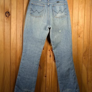 May include: Light blue denim jeans hanging on a wooden hanger. The jeans have a classic five-pocket design with the signature "W" stitching on the back pockets. The jeans are slightly faded, with a worn look.