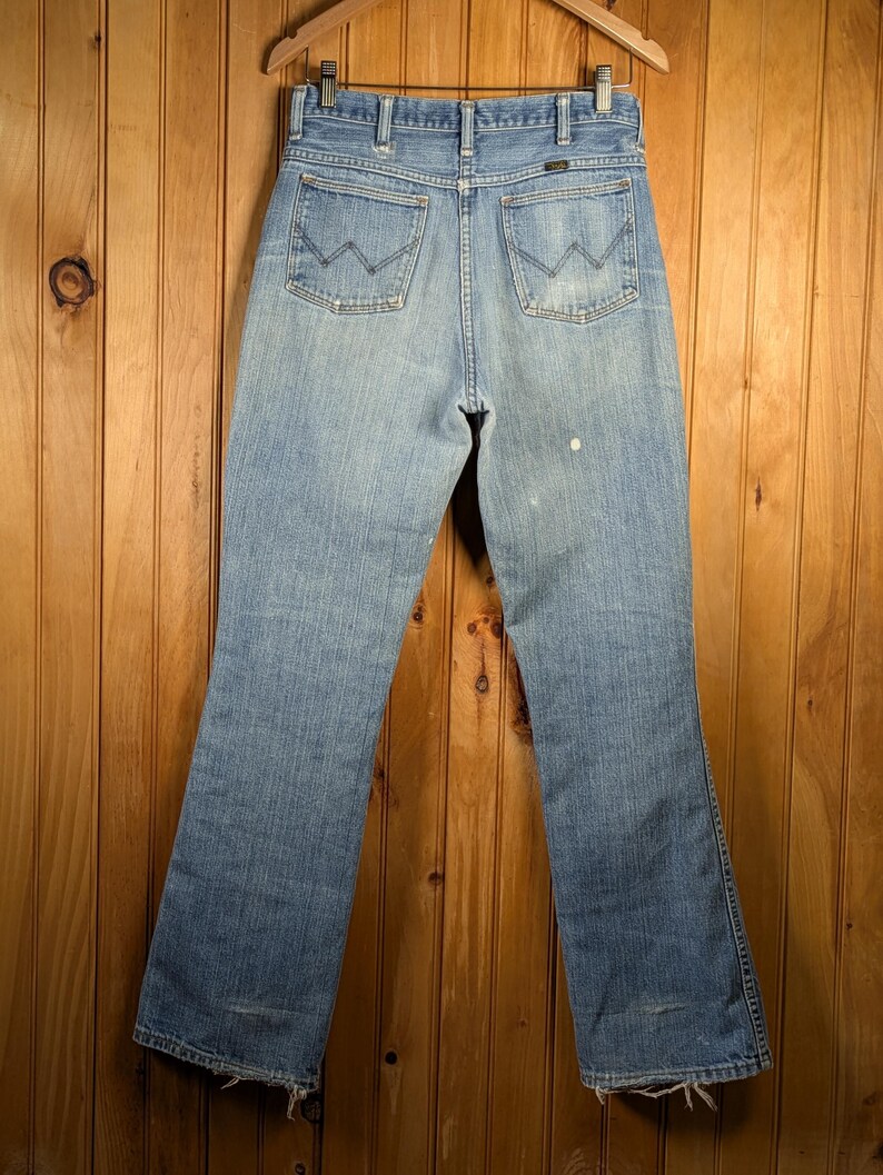 May include: Light blue denim jeans hanging on a wooden hanger. The jeans have a classic five-pocket design with the signature "W" stitching on the back pockets. The jeans are slightly faded, with a worn look.