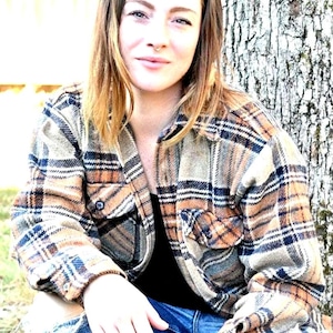 May include: A woman wearing a brown and black plaid flannel shirt with blue jeans. She is sitting in front of a tree trunk.