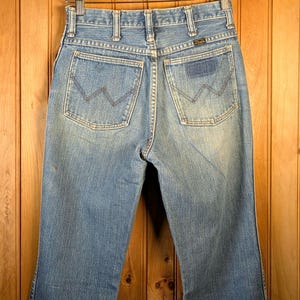 May include: Light blue denim jeans with a classic five-pocket design, featuring the Wrangler logo on the back pocket. The jeans are hanging on a wooden hanger against a wooden background. The jeans have a faded wash.