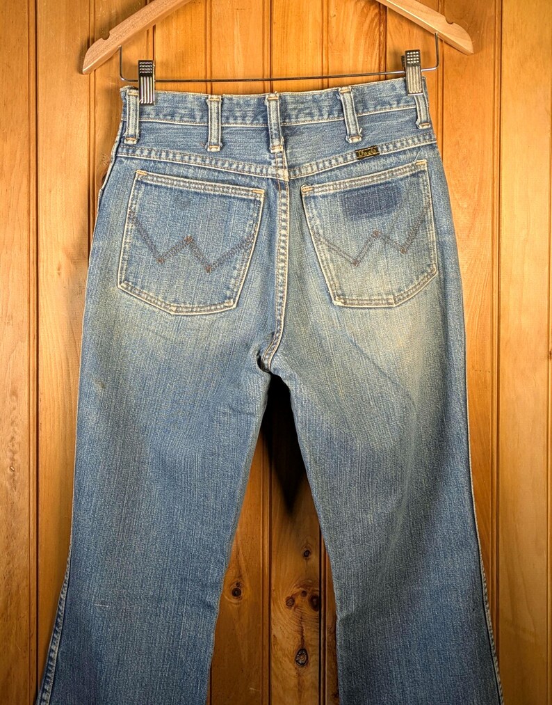 May include: Light blue denim jeans with a classic five-pocket design, featuring the Wrangler logo on the back pocket. The jeans are hanging on a wooden hanger against a wooden background. The jeans have a faded wash.