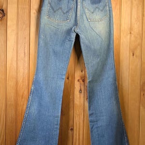 May include: Light blue denim jeans with a flared leg, hanging on a wooden hanger. The jeans have classic back pockets with a stitched design and a small logo patch. The jeans are displayed against a wooden panel background.