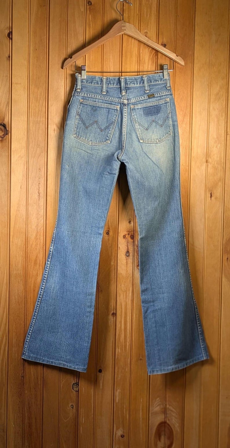 May include: Light blue denim jeans with a flared leg, hanging on a wooden hanger. The jeans have classic back pockets with a stitched design and a small logo patch. The jeans are displayed against a wooden panel background.