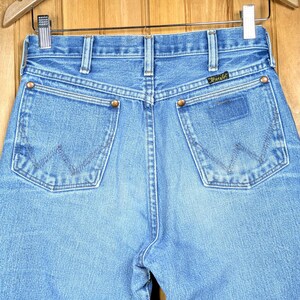 May include: Light blue denim jeans with the Wrangler logo on the back pocket. The jeans have classic five-pocket styling, copper rivets, and a straight leg. The back pockets feature the signature Wrangler "W" stitching.
