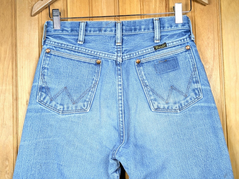 May include: Light blue denim jeans with the Wrangler logo on the back pocket. The jeans have classic five-pocket styling, copper rivets, and a straight leg. The back pockets feature the signature Wrangler "W" stitching.