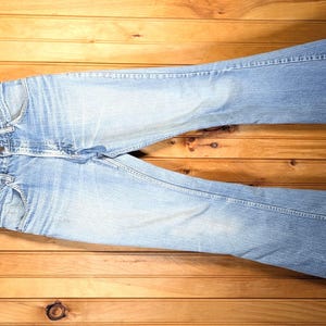 May include: Light blue denim jeans with a flared leg silhouette, featuring a classic five-pocket design, a button fly closure, and visible fading on the thighs and knees, hanging from a wooden hanger.