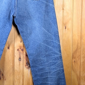 May include: Blue denim jeans with the Wrangler logo on the back pocket. The jeans are hanging on a wooden hanger against a wooden background. The jeans have a classic, straight-leg fit.