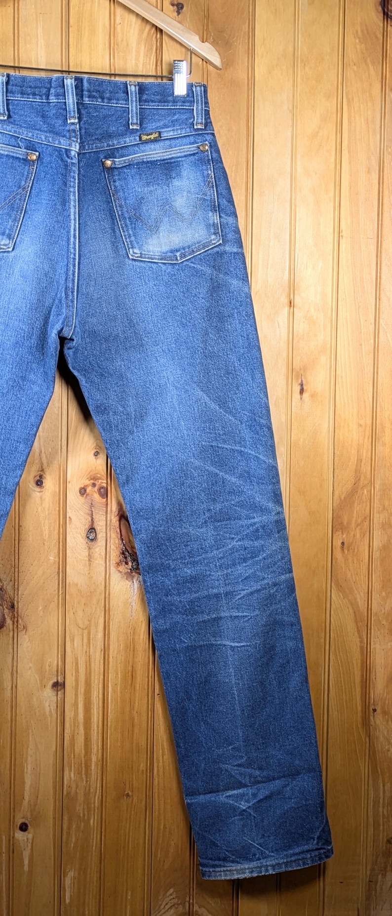 May include: Blue denim jeans with the Wrangler logo on the back pocket. The jeans are hanging on a wooden hanger against a wooden background. The jeans have a classic, straight-leg fit.