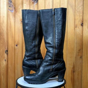 May include: A pair of black leather knee-high boots with gold zippers on the sides and a block heel.