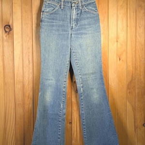 May include: Light blue denim jeans with a flared leg design, hanging on a wooden hanger. The jeans have a classic five-pocket design and a button and zipper closure. The denim has a faded appearance.
